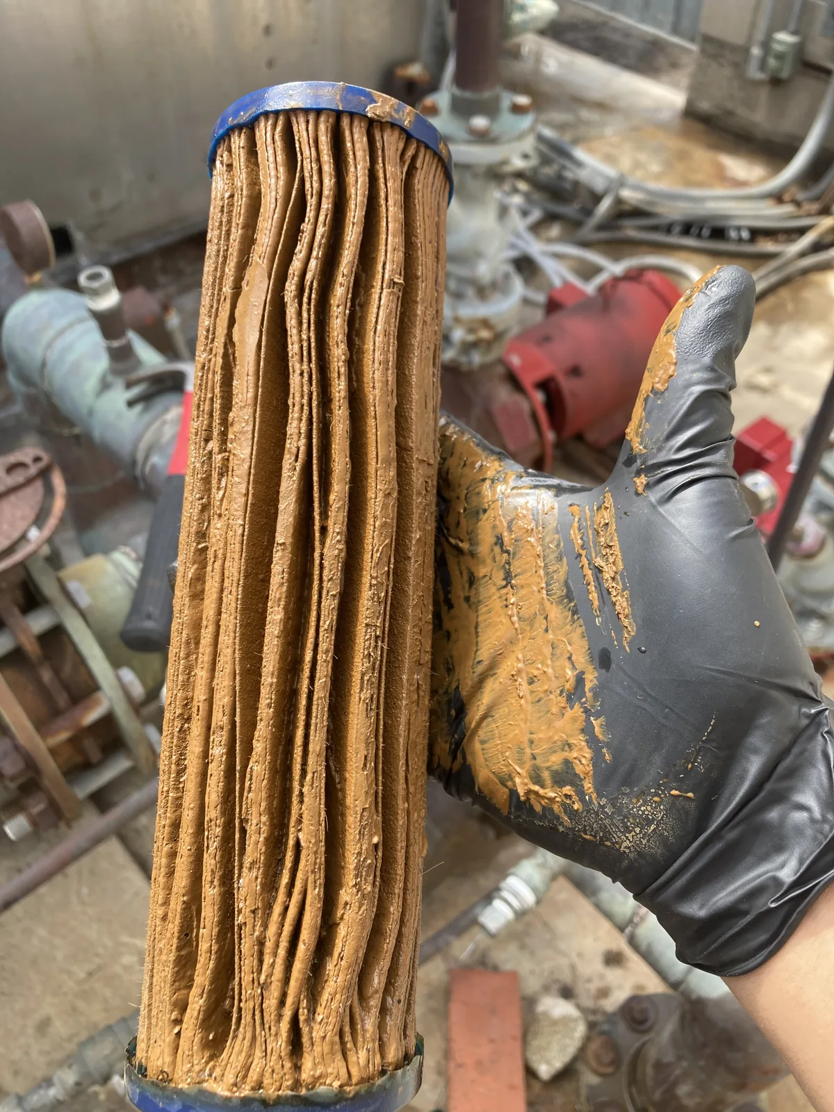 Pleated cartridge filter packed with orange-brown sludge, held in a gloved hand